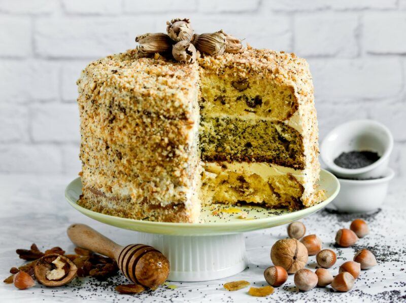 A white plate with a tall sour cream cake called smetannik, with many layers, with nuts scattered around the bottom and a honey stirrer