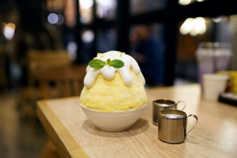 A wooden board with a bowl of yellow bird snow cone that has been topped with icing