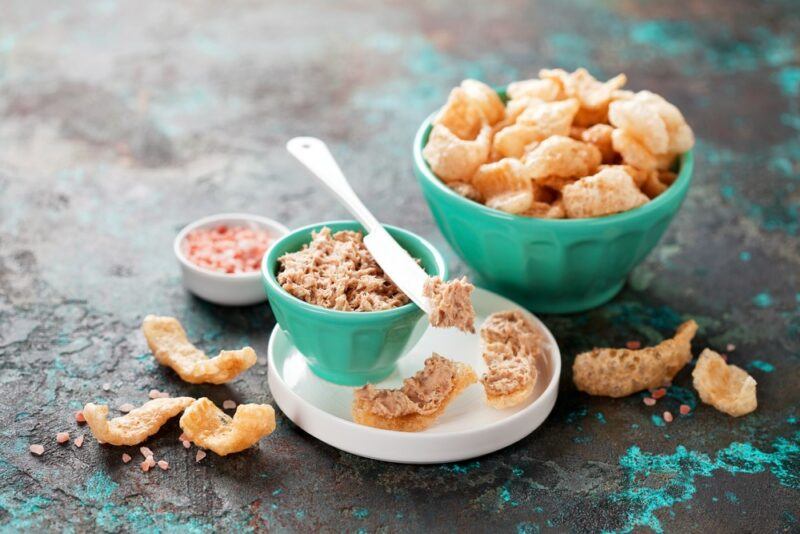 A large teal bowl containing pork rinds, next to a smaller bowl of dip and some more pork rinds