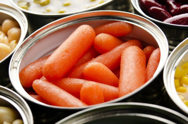 A tin containing cooked baby carrots, surrounded by partial tins containing other vegetables