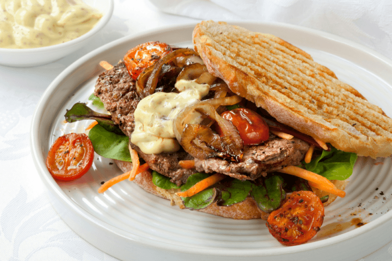 A large white plate containing a toasted steak sandwich with cheese and vegetables.