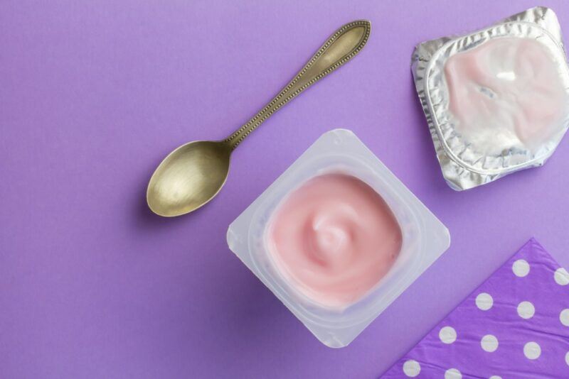 A top down image of a pottle of yogurt with pink yogurt, next to a lid, and a spoon