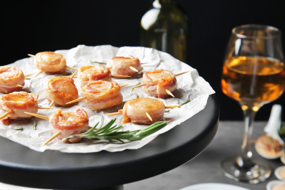 A platter with bacon wrapped scallops next to a glass of wine