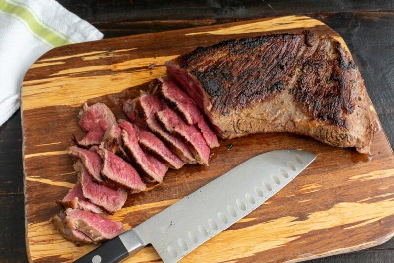 A large wooden board with a cooked tri tip steak that has been partially cut into small slices, next to a knife