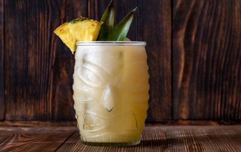 A tiki mug containing a tropical painkiller cocktail, garnished with a pineapple wedge and some pineapple leaves