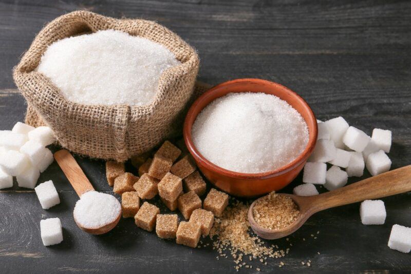 A wooden table with various types of sugar in a bag, a bowl, spoons, and cubes on the table