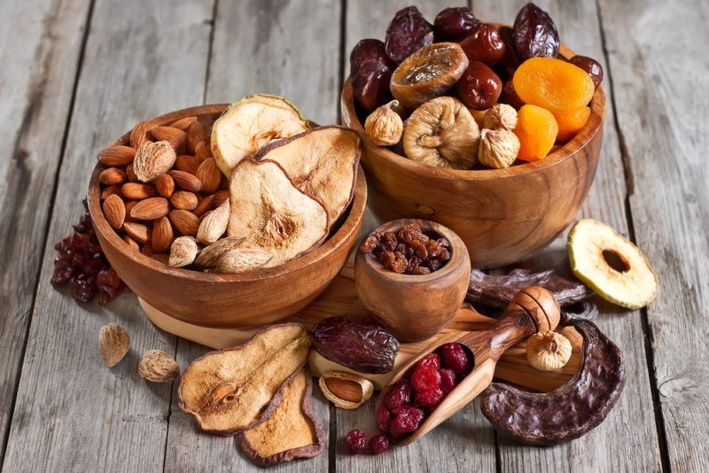 Three wooden bowls of various sizes filled with dried fruit and almonds, with more dried fruit on the table