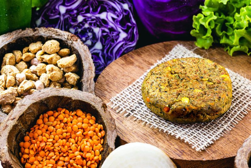 A vegan meat patty on a board with bowls of ingredients like lentils, nuts, and cabbage