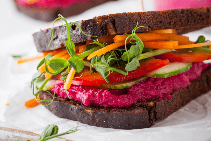 A large vegetable heavy sandwich that includes beet hummus, carrots, peppers, and greens.