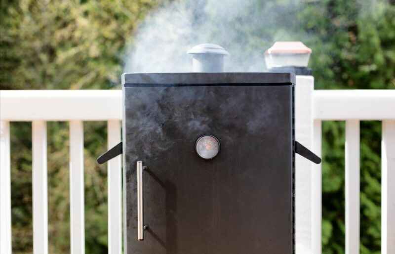 A vertical propane smoker outside by a railing | Food For Net A closed vertical smoker outside with smoke coming out the top