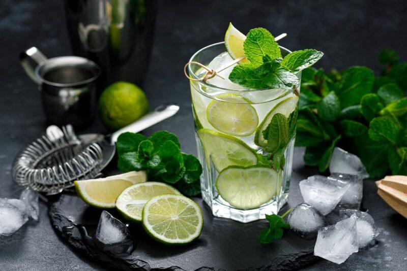 A tall glass containing a vodka mojito cocktail, surrounded by limes and ice