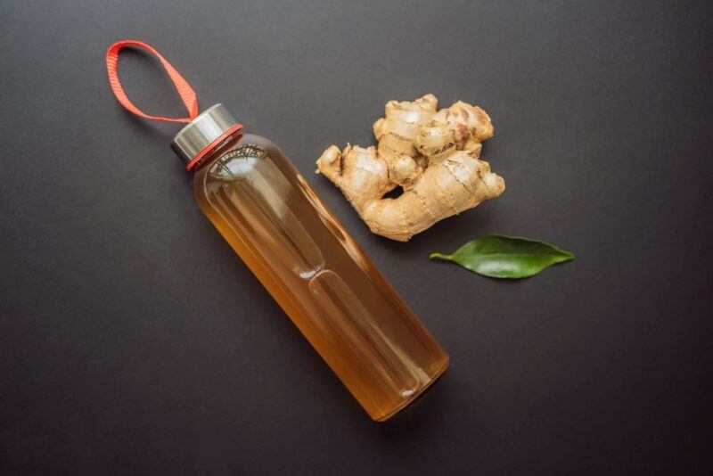 A black background with a glass water bottle filled with homemade ginger ale, next to some ginger root