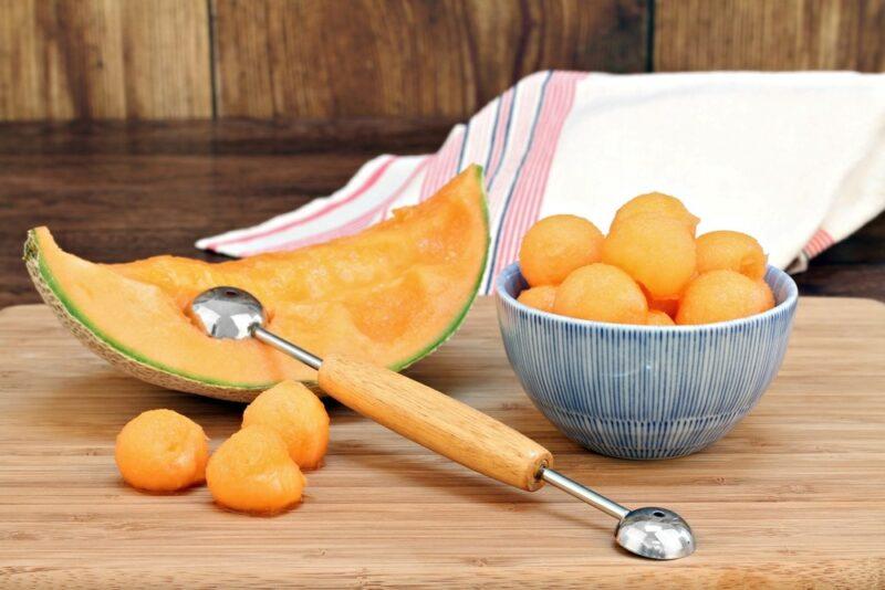 A wedge of cantaloupe with a melon baller and a bowl of cantaloupe balls