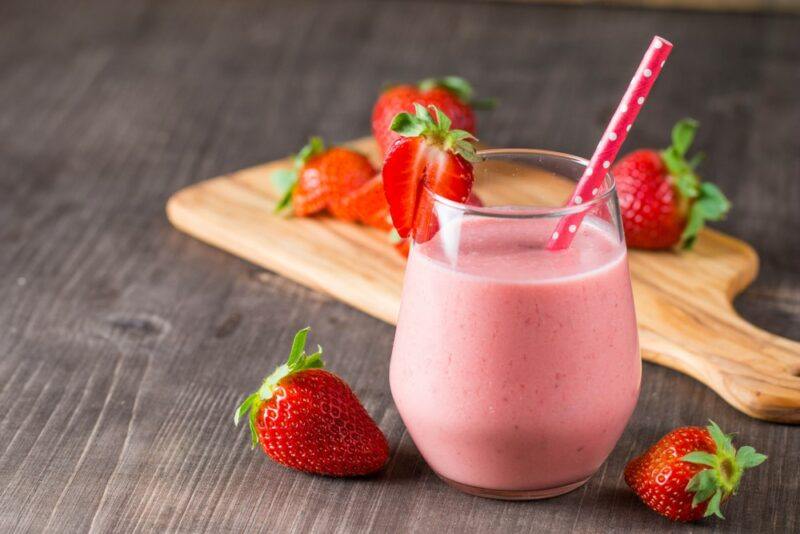 A glass containing a whiskey strawberry horchata cocktail, surrounded by strawberries, with a wooden board