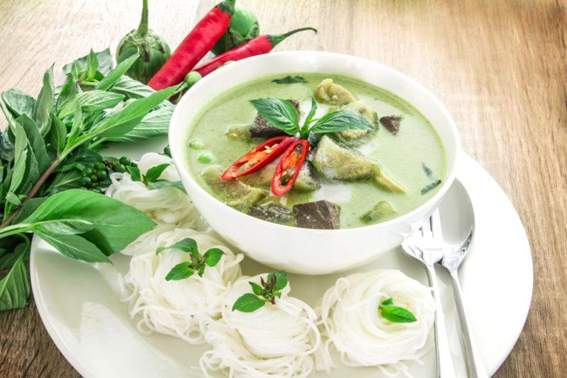 A white bowl of Thai green curry on a plate with rice or something similar, next to a leaf