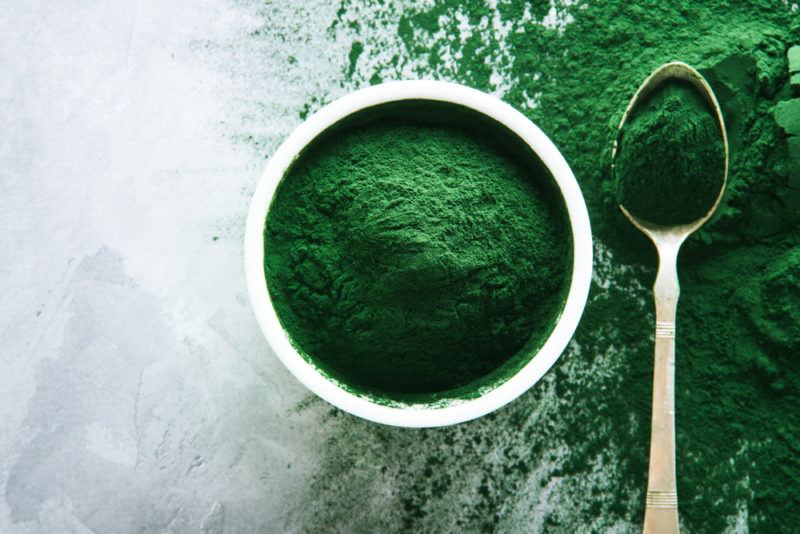 A white bowl and a spoon of spirulina, with spirulina scattered across the table