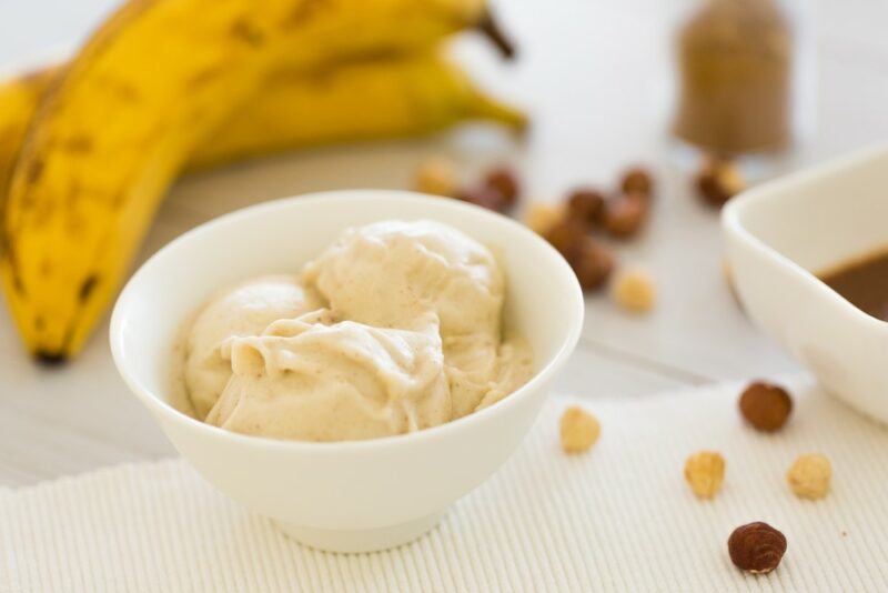 A small white bowl with banana ice cream (or nice cream), with some bananas in the background and a few toppings