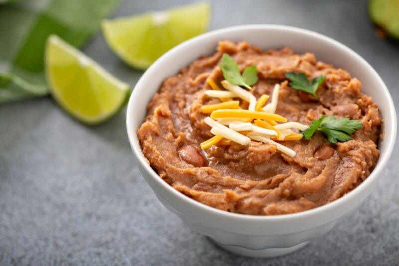 A white bowl of refried beans with cheese, with lime wedges in the background