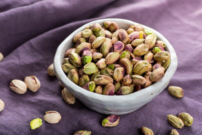 A white bowl containing shelled pistachio nuts on a purple cloth, with various pistachios scattered across the cloth