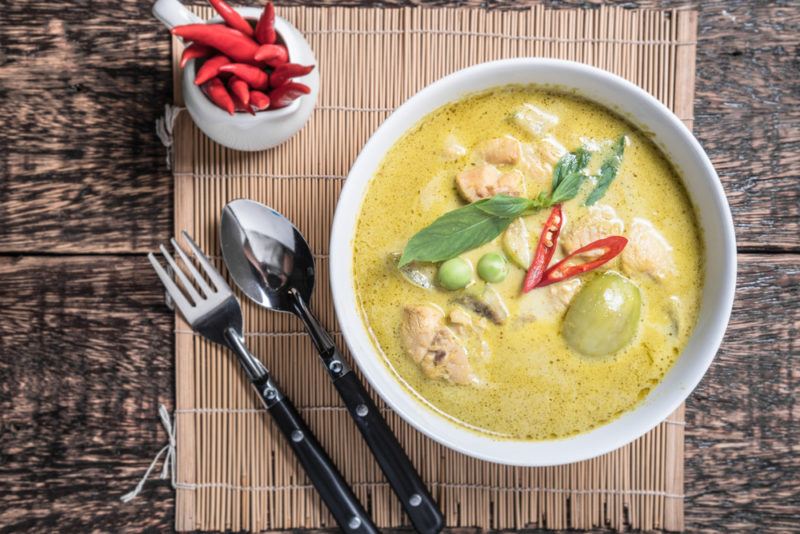 A white bowl of Thai green curry on a placemat, next to a knife and spoon, and spices