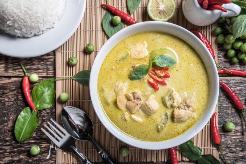 A white bowl of Thai green curry next to a plate of white rice and various spices on the table