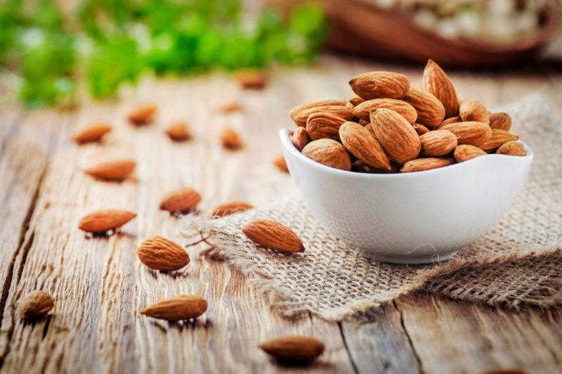 A small white bowl with almonds, on a wooden table with almonds scattered across it