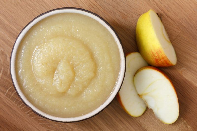 A wooden table with a bowl of apple sauce next to some apple slices