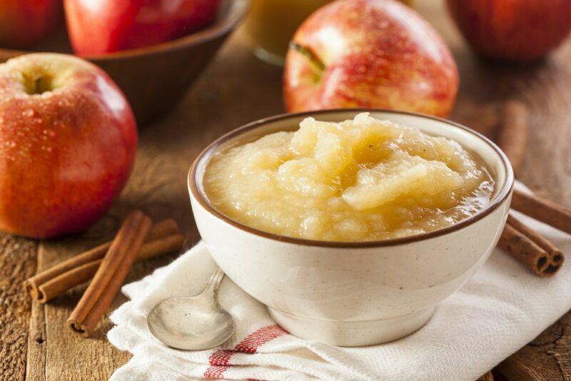 A white bowl containing apple sauce, with a few fresh apples and some cinnamon sticks