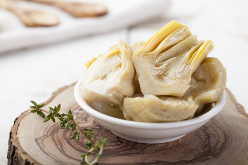 A white bowl of artichoke hearts on a wooden stump with a plate of spoons in the background