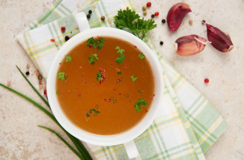 A top down view of a white bowl filled with bone broth, with seasoning ingredients scattered around