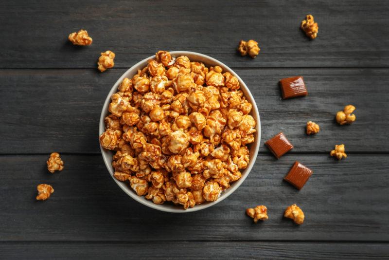 A bowl of caramel popcorn on a table, with a few pieces of popcorn and some caramel scattered on the table