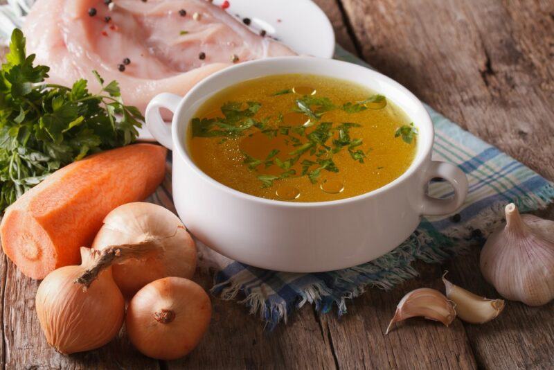 A wooden table with a bowl of chicken broth, next to onions, carrots, and some meat