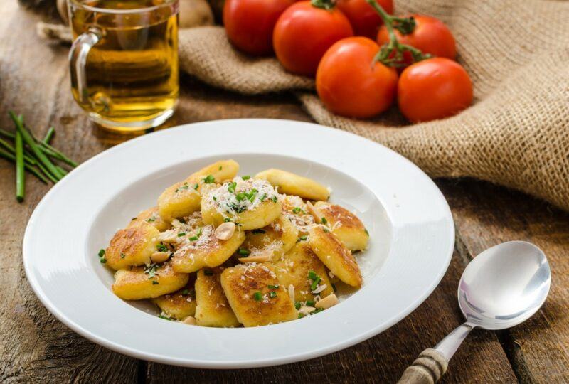 A white bowl of gnocchi on a wooden table next to a spoon, a jug of olive oil and some fresh tomatoes