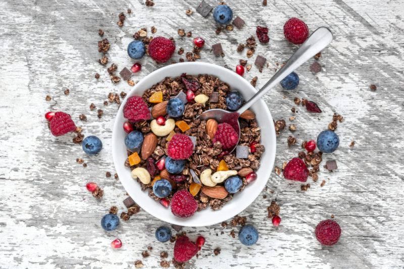 A white bowl of granola on a wooden table with fruit in the bowl and scattered around the table