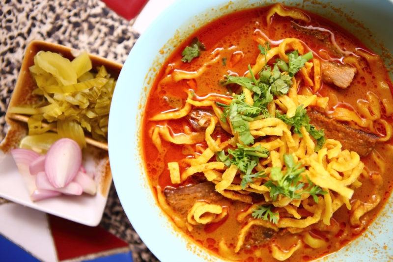 A white bowl with a red soup, called khao soi, and fried egg noodles