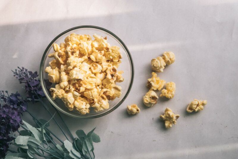 A glass bowl containing popcorn, with some on the table and some lavender