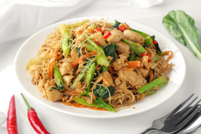 A white bowl containing a meal called mi xao, with some greens and chili peppers on the table