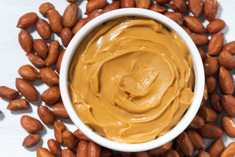 A white bowl of peanut butter with peanuts | Food For Net A white bowl of smooth peanut butter on top of a collection of peanuts in their skins