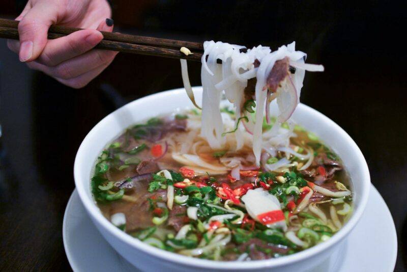 A large white bowl filled with pho, that someone is using chopsticks on