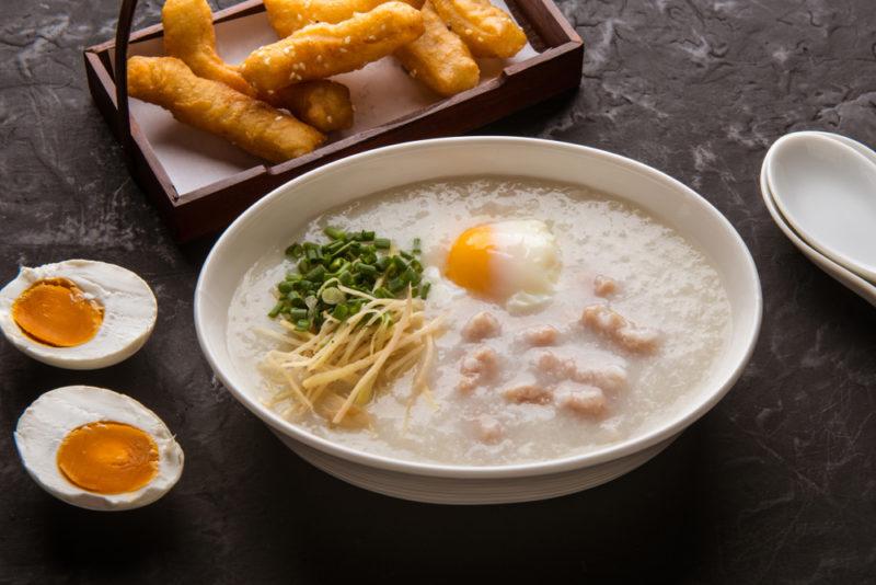 A white bowl with rice porridge, with eggs and fried dough sticks next to it