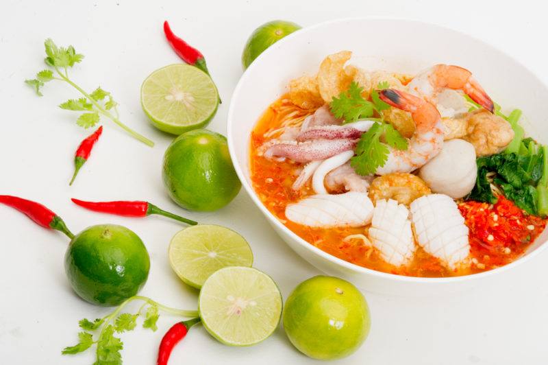 A white bowl that contains yum talay, next to peppers, limes and herbs