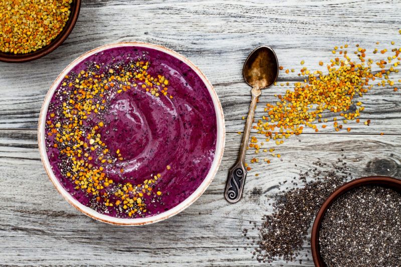 A white bowl with an acai smoothie and bee pollen. There is a spoon next to the bowl and bee pollen scattered across the table