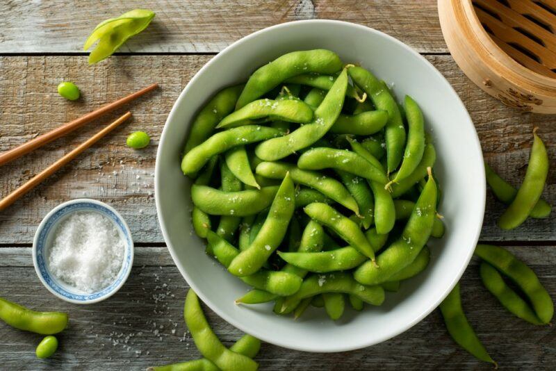 A large white bowl filled with cooked edamame, next to some chopsticks and a small container of salt