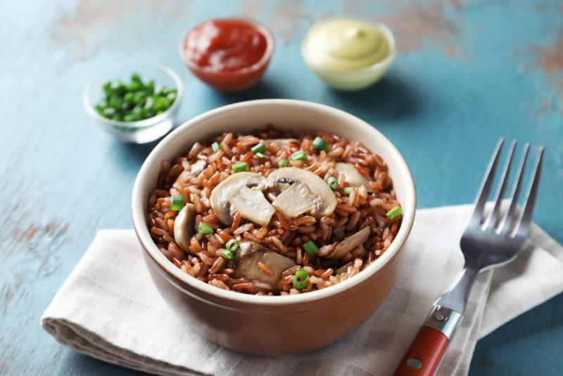 A brown bowl that contains cooked rice and mushrooms, with sauces in the background