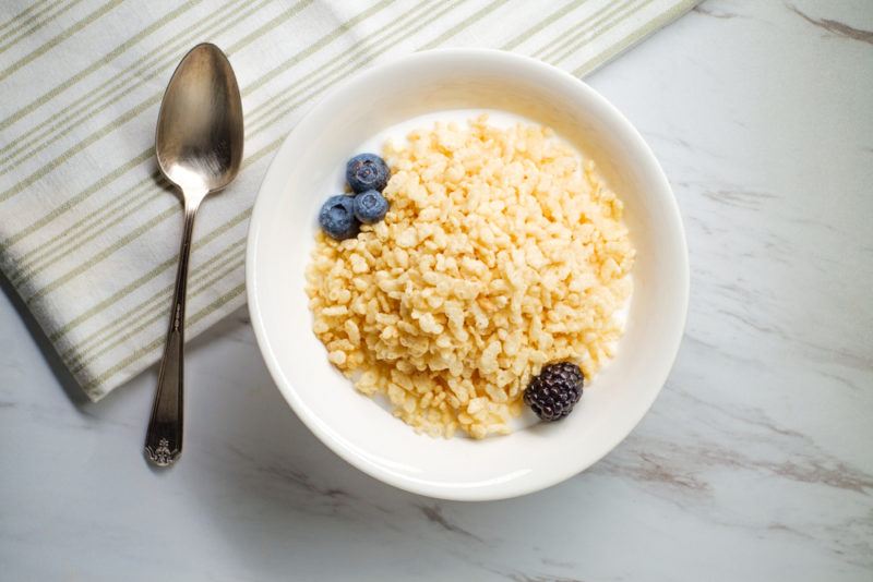 A white bowl that contains fortified breakfast cereal next to a stainless steel spoon