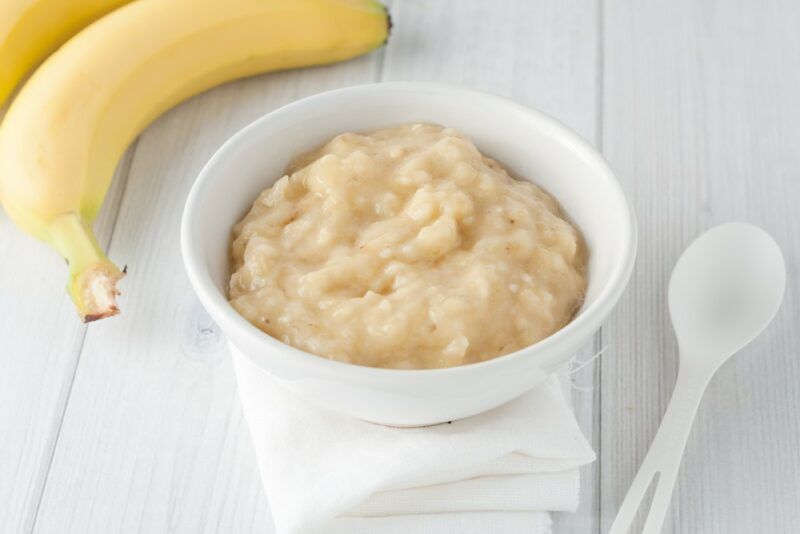 A white bowl filled with mashed banana next to a white spoon, with a couple of bananas in the background