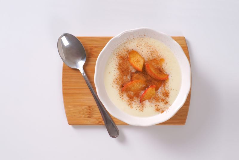 A white bowl and spoon on a small wooden board. The bowl contains a semolina pudding with fruit and cinnamon