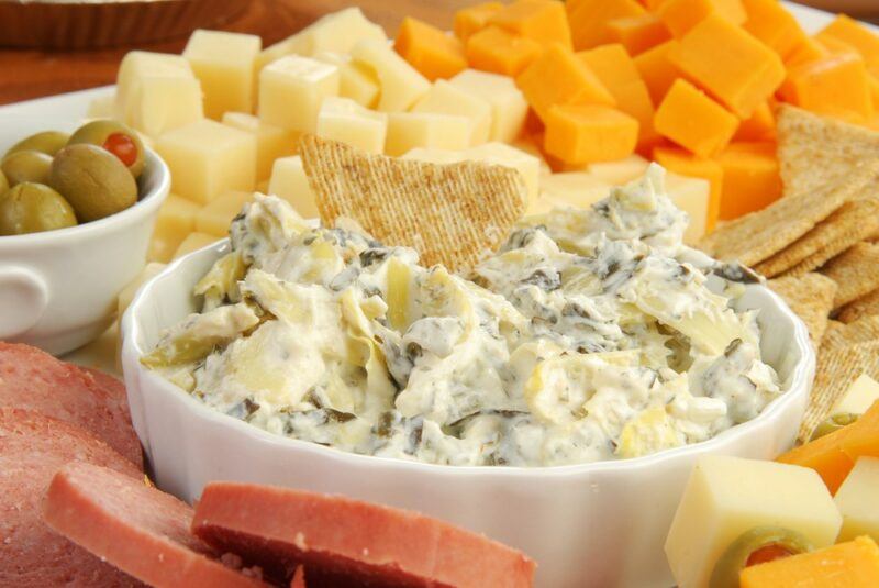 A large white container containing artichoke dip, with pieces of cheese and olives in the background