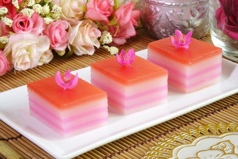 A white tray with three pieces of layered pink khanom chan dessert, with a selection of bright flowers in the background