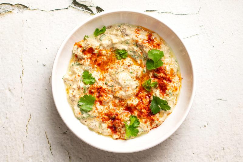 A white dish of baba ganoush with greens and a red spice